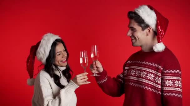 Portré fiatal pár karácsonyi Mikulás kalap iszik pezsgőt piros stúdió háttér. Szerelem, ünnepek, boldogság koncepció. — Stock videók