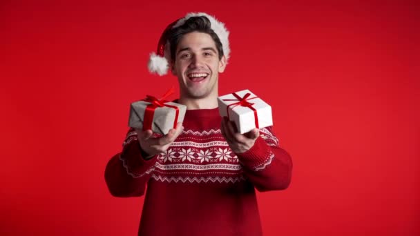 Ung stilig man ler och håller presentförpackning på röd studio bakgrund. Killen i tomteluva och ful jultröja. Nytt år humör. — Stockvideo