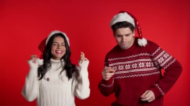 Kırmızı stüdyo arka planında Noel Baba şapkaları içinde dans eden genç bir çift. Aşk, tatiller, mutluluk konsepti. — Stok video
