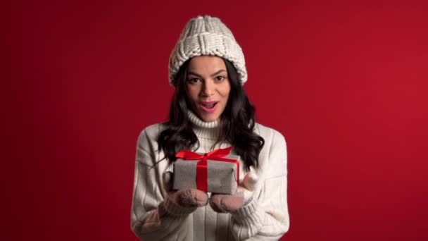 年轻可爱的女孩笑着拿着礼品盒,背景是红色的工作室. 戴着温暖针织帽子的女人. 新年气氛. — 图库视频影像