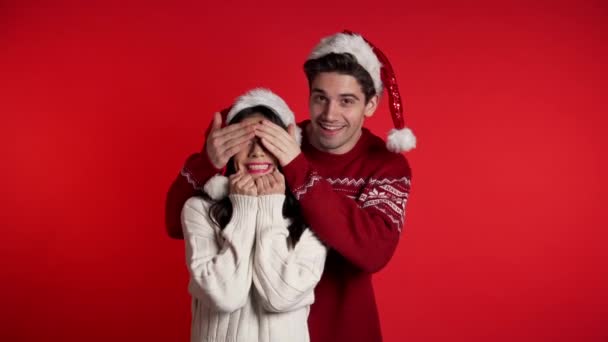 L'homme ferme les yeux de sa petite amie bien-aimée avant de la surprendre. Couple en chapeau de Père Noël sur fond de studio rouge. Amour, vacances, bonheur concept . — Video