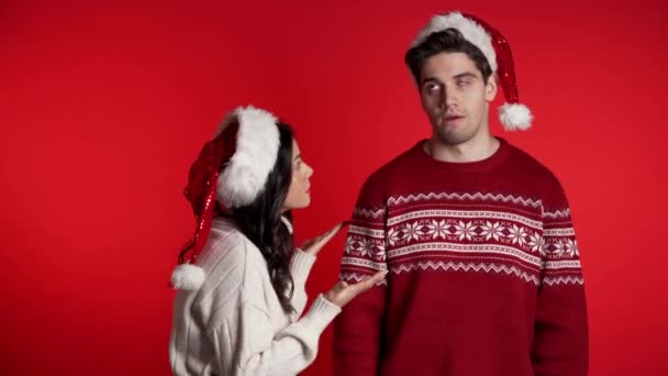 Young woman in Christmas hats emotionally scolds her boyfriend on red background in studio. Bored man rolling his eyes. Concept of conflict, problems in relationships. — ストック動画