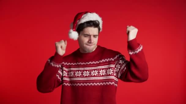 Hezký evropský muž v Santa klobouku a ošklivý svetr s úsměvem a tancem ve vynikající náladě na červeném pozadí. Vánoce, Silvestr, párty koncept. — Stock video