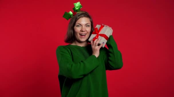 Młoda kobieta uśmiecha się i trzyma pudełko z prezentami w pobliżu ucha, żeby zgadnąć, co jest w środku na czerwonym tle studia. Dziewczyna w kapeluszu Mikołaja. Świąteczny nastrój. — Wideo stockowe