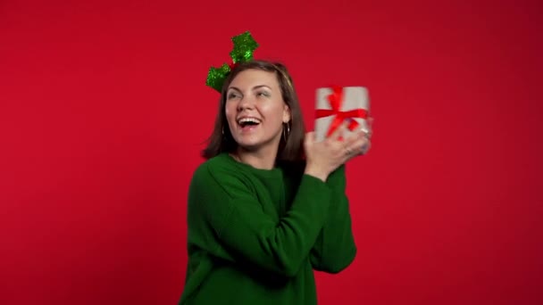 Mujer joven sonriendo y sosteniendo caja de regalo cerca de la oreja para adivinar lo que hay dentro en el fondo rojo del estudio. Una chica con sombrero de Santa. Humor de Navidad . — Vídeos de Stock