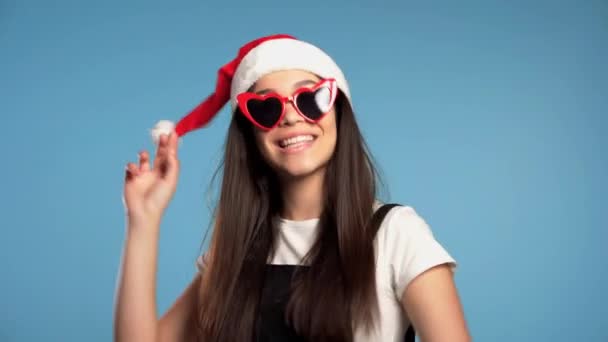 Attraktives Mädchen mit Weihnachtsmütze und herzförmiger Sonnenbrille, das Weihnachten oder Neujahr auf blauem Hintergrund feiert. glückliche hübsche Frau, die lächelt und Spaß hat. — Stockvideo