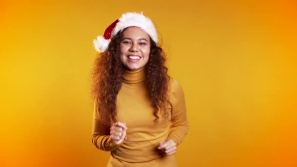 年轻可爱的女孩笑着在黄色工作室的背景上跳舞。 戴着圣诞礼帽的女人 新年气氛. — 图库视频影像