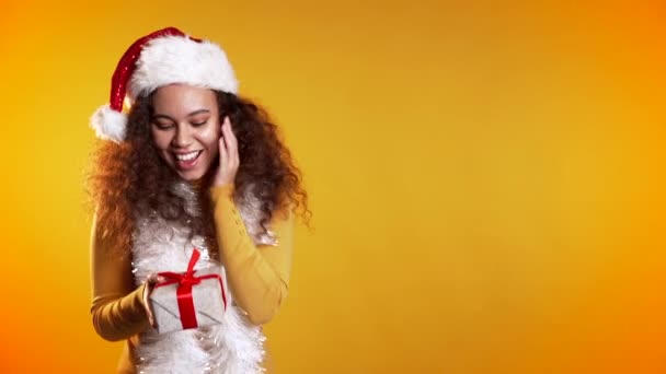 Surprised mixed race woman smiling and holding gift box on yellow studio background. Girl with curly hairstyle in sweater. Christmas mood. Copy space — ストック動画