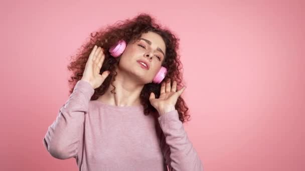 Hermosa mujer con pelo rizado bailando con auriculares sobre fondo de estudio rosa. Retrato de chicas lindas. Música, radio, felicidad, libertad, concepto de juventud . — Vídeos de Stock