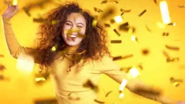 Izgatott lány táncol, tapsol, szórakozik, örül a konfetti esőnek a sárga stúdióban. Fogalom karácsony, újév, boldogság, party, győzelem — Stock videók