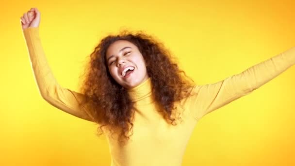 Menina bonito jovem sorrindo e dançando no fundo do estúdio amarelo. Mulher em desgaste brilhante colorido. Humor positivo. Movimento lento . — Vídeo de Stock