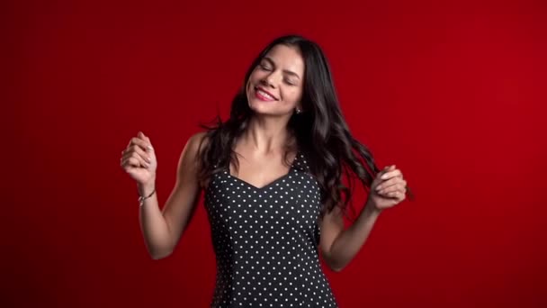 Atractiva mujer sexy sonriendo y bailando sobre fondo rojo estudio. Señora con el pelo largo en vestido de lunares de noche. Humor positivo. Movimiento lento — Vídeos de Stock