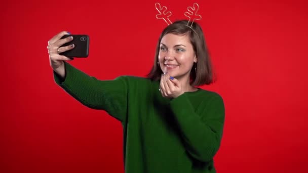 Mujer feliz sonriente en suéter verde y cuernos de ciervo haciendo selfie en el teléfono inteligente sobre fondo rojo colorido. Humor de Navidad. Movimiento lento — Vídeo de stock