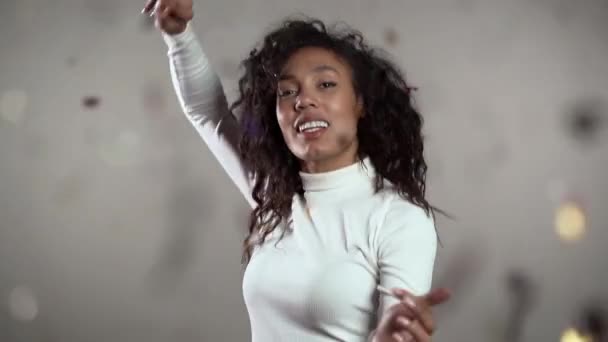 Femme afro-américaine excitée dansant, s'amusant, se réjouit de la pluie de confettis en studio gris. Concept de Noël, Nouvel An, bonheur, fête, gagner — Video