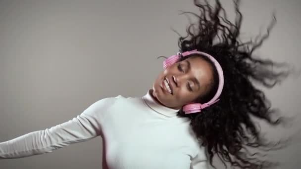 Mujer afroamericana con pelo rizado divirtiéndose, sonriendo, bailando con auriculares en estudio sobre fondo gris. Música, danza, concepto de radio, cámara lenta . — Vídeo de stock
