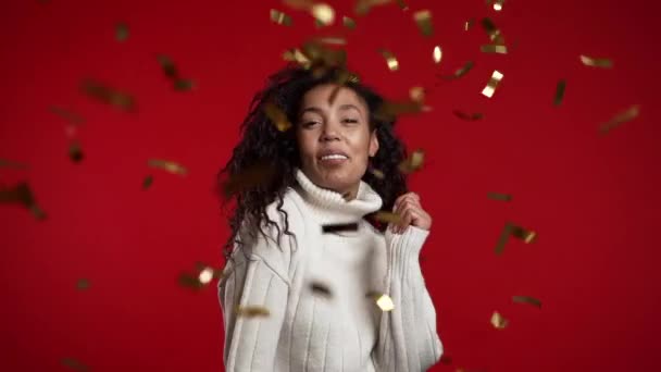 Mulher afro-americana excitada dançando, se divertindo, se alegra com a chuva de confetes no estúdio vermelho. Conceito de Natal, Ano Novo, felicidade, festa, vitória — Vídeo de Stock