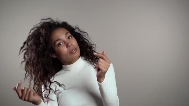 Mulher afro-americana com cabelo encaracolado se divertindo, sorrindo, dançando com a cabeça e estala os dedos em estúdio contra fundo cinza. Música, conceito de dança, câmera lenta . — Vídeo de Stock