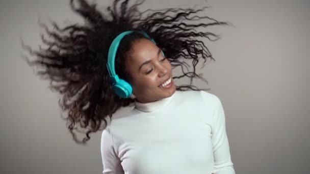Αφροαμερικανή γυναίκα με σγουρά μαλλιά που διασκεδάζει, χαμογελά, χορεύει με ακουστικά στο στούντιο με γκρι φόντο. Μουσική, χορός, ραδιοφωνική ιδέα, αργή κίνηση. — Αρχείο Βίντεο