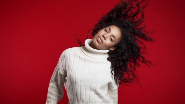 Mulher afro-americana com cabelo encaracolado se divertindo, sorrindo, dançando com a cabeça em estúdio contra fundo vermelho. Música, conceito de dança, câmera lenta . — Vídeo de Stock