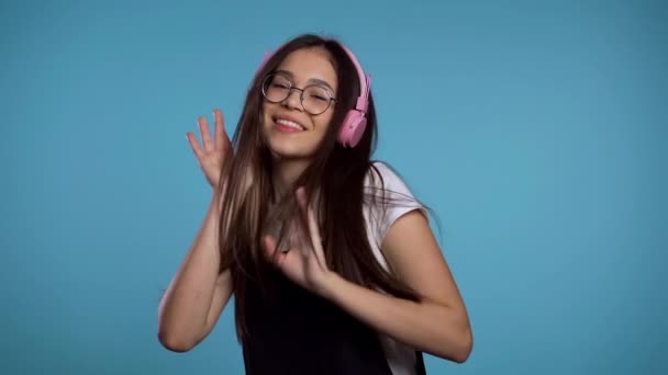 Hezká mladá asijská dívka s dlouhými vlasy baví, usmívá se, tančí se sluchátky ve studiu na modrém pozadí. Hudba, tanec, koncept rádia, zpomalení — Stock video