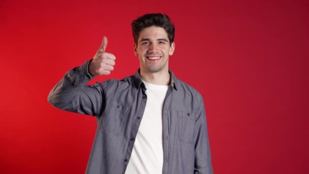 El joven positivo sonríe a la cámara. Hipster tipo mostrando el pulgar hacia arriba signo sobre fondo rojo. Ganador. Éxito. Lenguaje corporal . — Vídeos de Stock