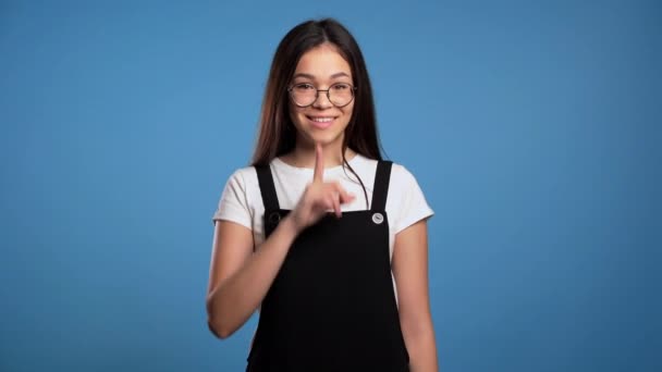 Glimlachende Aziatische vrouw met lang haar met vinger op haar lippen over een blauwe achtergrond. Het gebaar van de stilte, het geheim. Sluiten.. — Stockvideo