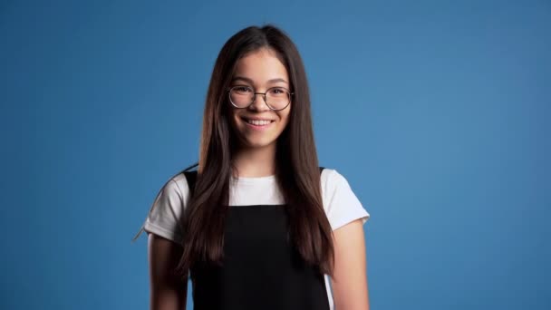 Hübsche junge asiatische Mädchen mit langen Haaren steht auf blauem Studiohintergrund. Niedliches Porträt einer Frau mit durchsichtiger runder Brille und schwarzem Overall. — Stockvideo