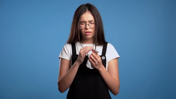 Young asian girl sneezes into tissue. Isolated woman on blue studio background is sick, has a cold or allergic reaction. Health, medicine, illness, treatment concept — Stock Video