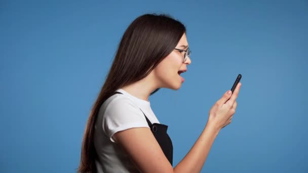 Boos Aziatische vrouw schreeuwt neer, zweert met iemand. Mobiele telefoon gebruiken. Gestresst en depressief meisje op blauwe achtergrond. — Stockvideo