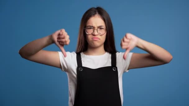 Jong Aziatisch meisje staat op blauwe studio achtergrond uiten ontevredenheid en tonen duim naar beneden gebaar op camera. Portret van een vrouw met tekenen van afkeer. — Stockvideo