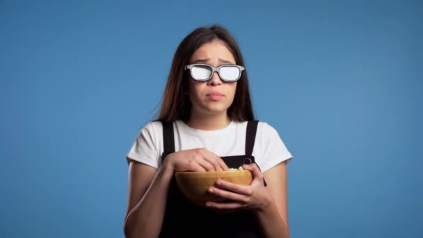 Joven mujer asiática en gafas 3d viendo películas de terror y comiendo palomitas de maíz sobre fondo de estudio azul. Asustada chica debido a la inesperada trama en el cine . — Vídeos de Stock