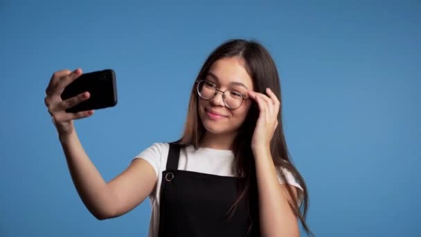 Lächelnd glückliches asiatisches Mädchen mit Brille macht Selfie auf Smartphone vor blauem Hintergrund. Technologie, Mobilgeräte, Konzept der sozialen Netzwerke. — Stockvideo