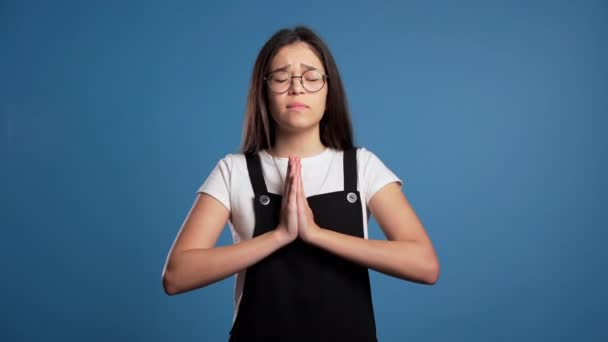 Mignon asiatique jeune fille priant sur fond bleu. Femme à lunettes mendiant quelqu'un. 4k . — Video