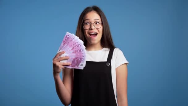 Ικανοποιημένο χαρούμενο ενθουσιασμένο κορίτσι από την Ασία που δείχνει χρήματα - χαρτονομίσματα του ευρώ στον μπλε τοίχο. Σύμβολο επιτυχίας, κέρδους, νίκης. — Αρχείο Βίντεο