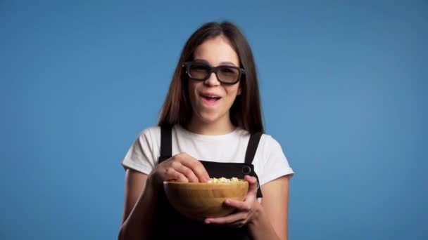 Ung asiatisk flicka i 3D-glasögon titta fascinerande komedi film, skrattar och äter popcorn på blå studio bakgrund. — Stockvideo