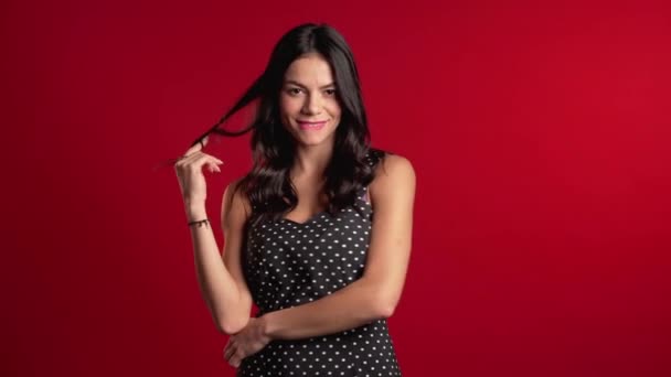 Красивая молодая латиноамериканка с длинными волосами, стоящая на красном фоне студии, улыбающаяся и флиртующая. Симпатичный портрет женщины в винтажном платье в горошек — стоковое видео
