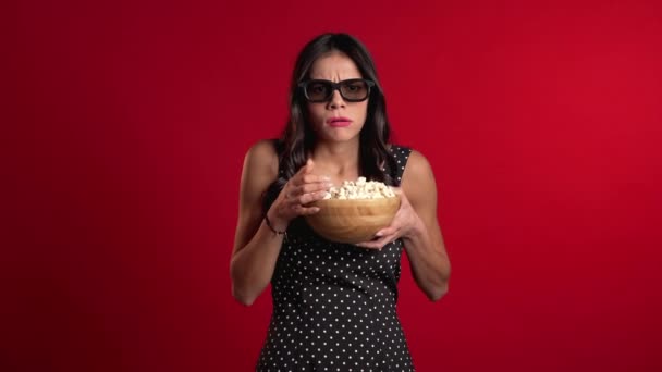 Mujer hispana joven en gafas 3D viendo películas de terror y comiendo palomitas de maíz en el fondo rojo del estudio. Asustada chica debido a la inesperada trama en el cine . — Vídeo de stock