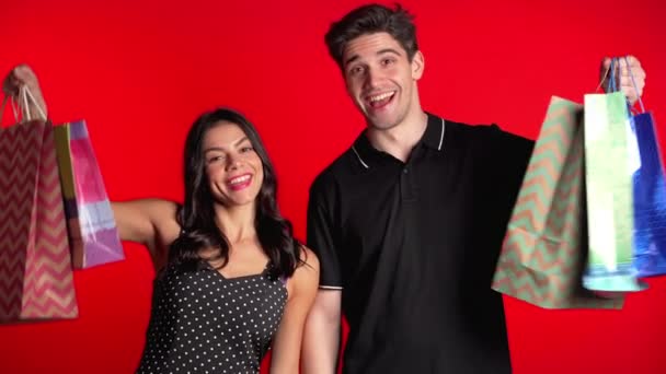 Feliz pareja joven con coloridas bolsas de papel después de comprar aislado en el fondo del estudio rojo. Venta estacional, compras, gasto en concepto de regalos — Vídeo de stock