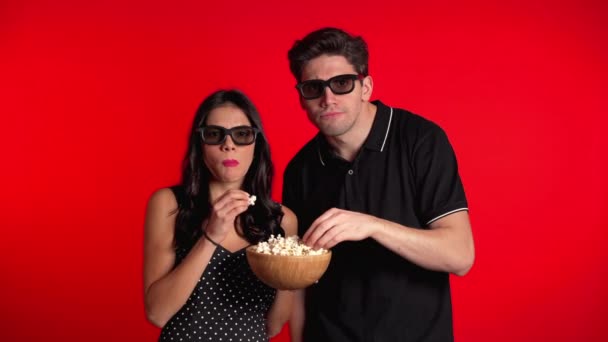 Young couple in cinema in 3d glasses watching fascinating horror movie and eating popcorn on red studio background. — Stock Video