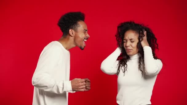 Mladý africký muž emocionálně křičí na svou ženu nebo přítelkyni na červeném pozadí ve studiu. Nudná žena zakryje uši rukama. Koncept konfliktu, problémy ve vztazích. — Stock video