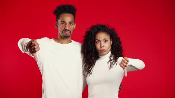 Unga afrikanska par står på röd studio bakgrund uttrycker missnöje och visar tummen ner gest vid kameran. Porträtt av man och kvinna med tecken på ogillande. — Stockvideo