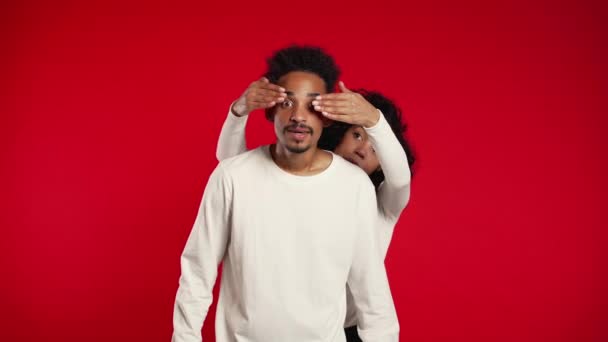 Afrikansk kvinna blundar för sin älskade pojkvän innan hon överraskar honom. Par i vitt på röd studio bakgrund. Kärlek, semester, lycka koncept. — Stockvideo