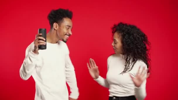 Femme africaine aux cheveux bouclés et homme souriant, dansant avec haut-parleur portable sans fil en studio sur fond rouge. Musique, concept de danse . — Video