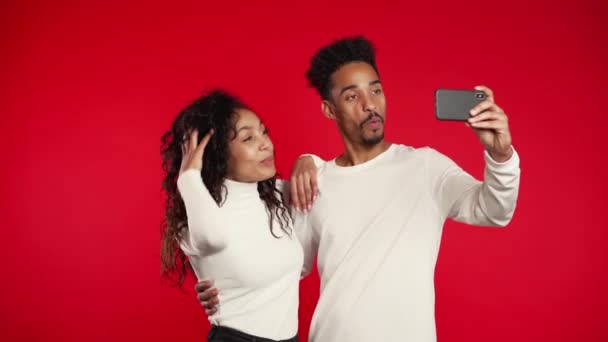 若いアフリカ系アメリカ人のカップルは、赤いスタジオの背景にスマートフォンで自撮りをしています。愛、休日、幸福の概念. — ストック動画
