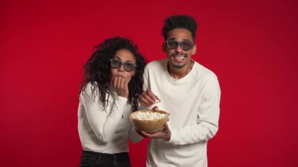 Joven pareja africana en el cine en gafas 3d viendo fascinante película de terror y comer palomitas de maíz en el fondo del estudio rojo . — Vídeos de Stock