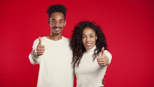 A fiatal afro-amerikai házaspár felemeli a hüvelykujját, piros háttérrel. Győztes. Sikeres volt. Pozitív lány és férfi mosolyog a kamerába. Testbeszéd. — Stock videók