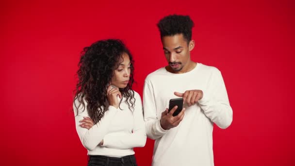 Молодая пара использует мобильное устройство. Мужчина рассказывает о приложениях и объясняет девушке, как пользоваться приложением. Красный фон студии — стоковое видео
