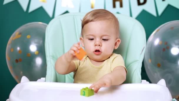 かわいい赤ちゃんは椅子に座ってカラフルなレンガで遊んでいます。大きな目の肖像画と誕生日ハンサムな幼児の子供。面白い子だ. — ストック動画