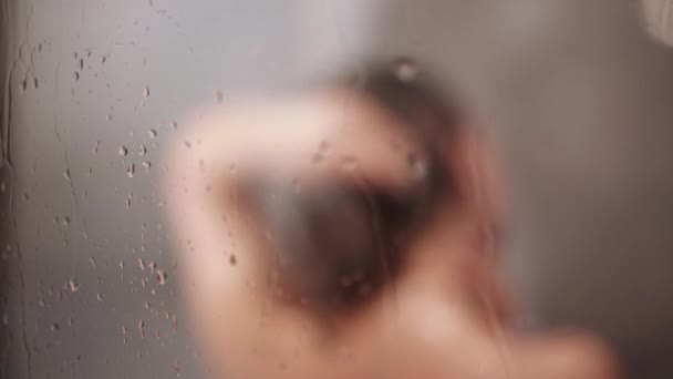 Naked woman washing brown hair with shampoo and body with gel in shower. Beauty care concept, mask. Focus on bath glass wall. Girl in defocus. — ストック動画