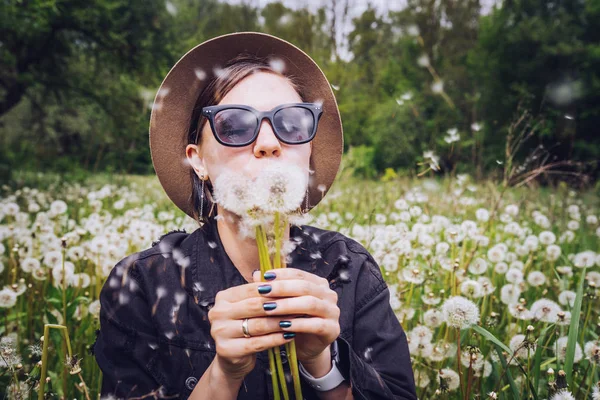 Mutlu kadın parkta karahindiba üfleme. Şapkalı ve güneş gözlüklüğü olan kız. Dilek, sevinç kavramı